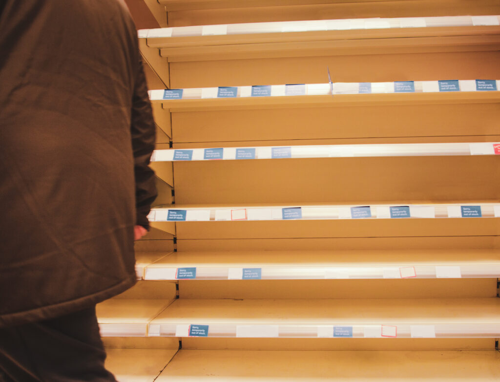 A figure walks past a set of bare pharmacy shelves. The impact of supply chain shortages can have a long tail.