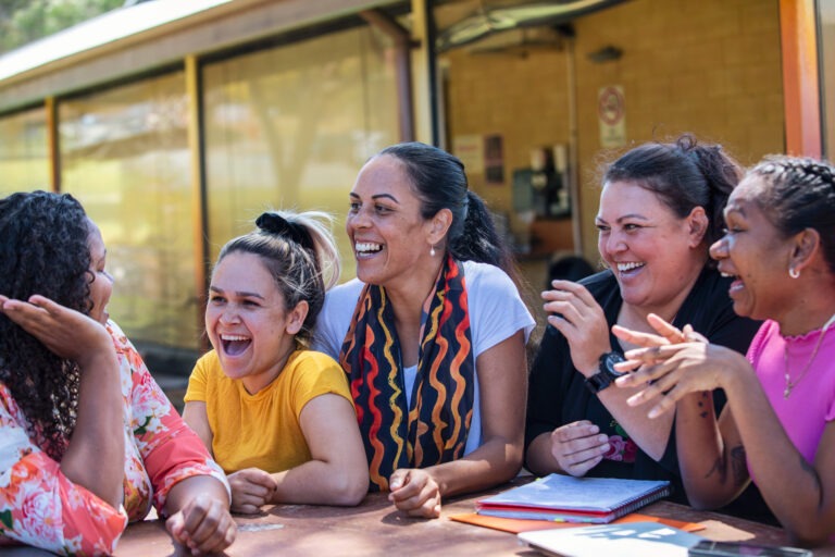 A group of Indigenous Australian women talking around a table. Aboriginal and Torres Strait Islander community-controlled health organisations take a vastly different approach from the mainstream health system.