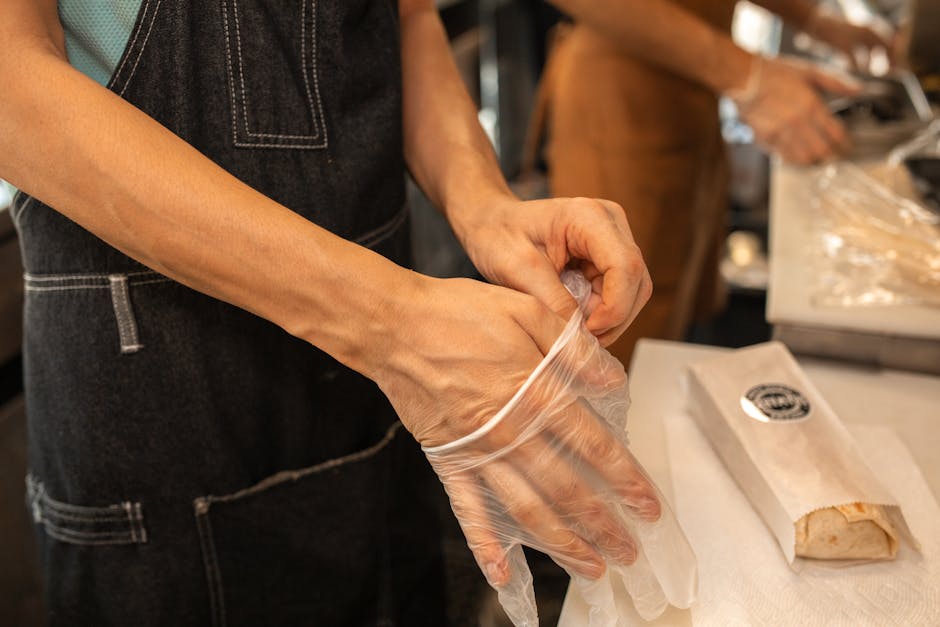 A person wearing an apron puts on plastic gloves in a kitchen environment, preparing for food handling.